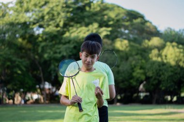 Kamu parkının çimlerinde arkadaşlarıyla dışarıda badminton oynamak için elinde beyaz pipi ve badminton raketi tutan Asyalı çocuk. Yumuşak ve seçici bir odak noktası..