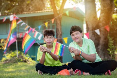 Asyalı oğlanlar gökkuşağı bayrağı, LGBT sembolü tutarlar, evlerinin önündeki çimenlikte otururlar. Evlerinin önündeki ağaçlarda asılı gökkuşağı teli bayrakları ile süslenmişlerdir..