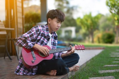 Asyalı tatlı çocuk balkonun cam kapısının önünde oturur ve akustik gitar çalar, yumuşak ve seçici odak, gün batımının ışıklarıyla düzenlenmiş arka plan.