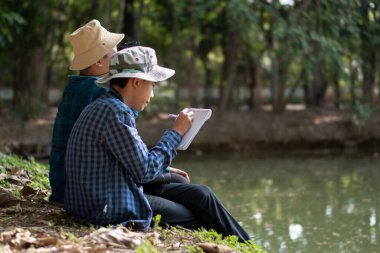 Asyalı çocuklar göletin kenarında oturmuş, ellerinde dürbünle kuşları izliyorlar ve doğa ile çevreyi birlikte öğreniyorlar..