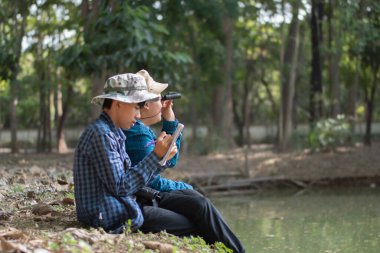 Asyalı çocuklar göletin kenarında oturmuş, ellerinde dürbünle kuşları izliyorlar ve doğa ile çevreyi birlikte öğreniyorlar..