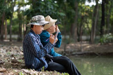 Asyalı çocuklar göletin kenarında oturmuş, ellerinde dürbünle kuşları izliyorlar ve doğa ile çevreyi birlikte öğreniyorlar..