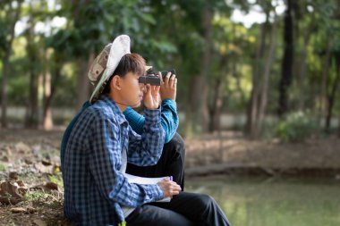 Asyalı çocuklar göletin kenarında oturmuş, ellerinde dürbünle kuşları izliyorlar ve doğa ile çevreyi birlikte öğreniyorlar..