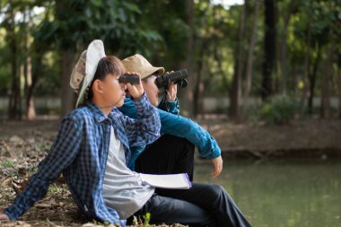 Asyalı çocuklar göletin kenarında oturmuş, ellerinde dürbünle kuşları izliyorlar ve doğa ile çevreyi birlikte öğreniyorlar..