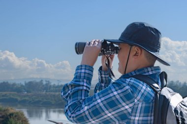 Asyalı bir çocuk, kep ve ekose gömlek giyiyordu. Dürbünle nehrin üzerindeki köprüde dikilip bakıyordu. Doğal ortamın tadını çıkarıyor ve yabani hayatı merakla izliyor..