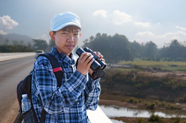 Asyalı bir çocuk, kep ve ekose gömlek giyiyordu. Dürbünle nehrin üzerindeki köprüde dikilip bakıyordu. Doğal ortamın tadını çıkarıyor ve yabani hayatı merakla izliyor..
