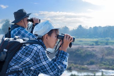 Asyalı bir çocuk, kep ve ekose gömlek giyiyordu. Dürbünle nehrin üzerindeki köprüde dikilip bakıyordu. Doğal ortamın tadını çıkarıyor ve yabani hayatı merakla izliyor..