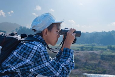 Asyalı bir çocuk, kep ve ekose gömlek giyiyordu. Dürbünle nehrin üzerindeki köprüde dikilip bakıyordu. Doğal ortamın tadını çıkarıyor ve yabani hayatı merakla izliyor..