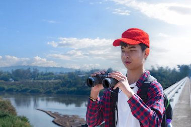 Asyalı bir çocuk, kep ve ekose gömlek giyiyordu. Dürbünle nehrin üzerindeki köprüde dikilip bakıyordu. Doğal ortamın tadını çıkarıyor ve yabani hayatı merakla izliyor..