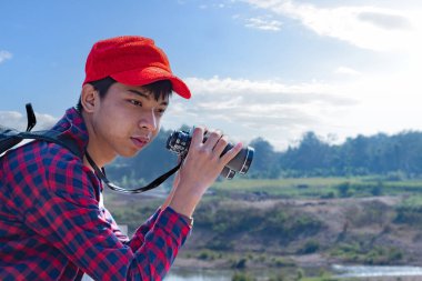 Asyalı bir çocuk, kep ve ekose gömlek giyiyordu. Dürbünle nehrin üzerindeki köprüde dikilip bakıyordu. Doğal ortamın tadını çıkarıyor ve yabani hayatı merakla izliyor..