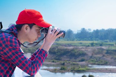 Asyalı bir çocuk, kep ve ekose gömlek giyiyordu. Dürbünle nehrin üzerindeki köprüde dikilip bakıyordu. Doğal ortamın tadını çıkarıyor ve yabani hayatı merakla izliyor..
