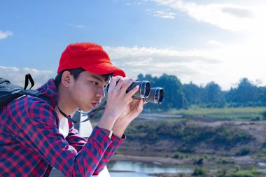 Asyalı bir çocuk, kep ve ekose gömlek giyiyordu. Dürbünle nehrin üzerindeki köprüde dikilip bakıyordu. Doğal ortamın tadını çıkarıyor ve yabani hayatı merakla izliyor..