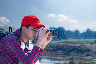 Asyalı bir çocuk, kep ve ekose gömlek giyiyordu. Dürbünle nehrin üzerindeki köprüde dikilip bakıyordu. Doğal ortamın tadını çıkarıyor ve yabani hayatı merakla izliyor..