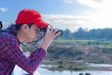 Asyalı bir çocuk, kep ve ekose gömlek giyiyordu. Dürbünle nehrin üzerindeki köprüde dikilip bakıyordu. Doğal ortamın tadını çıkarıyor ve yabani hayatı merakla izliyor..