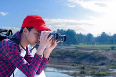 Asyalı bir çocuk, kep ve ekose gömlek giyiyordu. Dürbünle nehrin üzerindeki köprüde dikilip bakıyordu. Doğal ortamın tadını çıkarıyor ve yabani hayatı merakla izliyor..