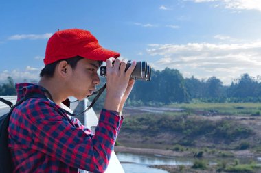 Asyalı bir çocuk, kep ve ekose gömlek giyiyordu. Dürbünle nehrin üzerindeki köprüde dikilip bakıyordu. Doğal ortamın tadını çıkarıyor ve yabani hayatı merakla izliyor..
