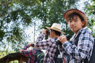 Asyalı çocuklar grup halinde durup dürbünle bakıyorlar ve parkta vahşi hayvan görmek için bekliyorlar. Doğal ortamın tadını çıkarıyorlar ve yabani hayatı merakla izliyorlar..