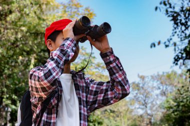 Dürbün tutan Asyalı çocuk tropikal ormanda duruyor ve parkta vahşi hayvan görmek için bekliyor. Doğal ortamın tadını çıkarıyor ve yabani hayatı merakla izliyor..