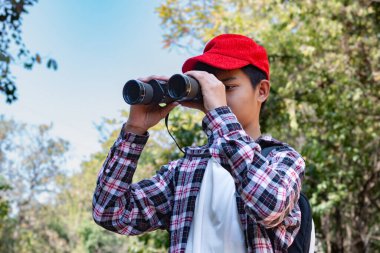 Asian boy holding binoculars and  standing in tropical forest waiting to see wildlife animals in local park. He is enjoying the natural surroundings and observing wildlife with curiosity. clipart