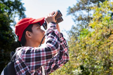 Dürbün tutan Asyalı çocuk tropikal ormanda duruyor ve parkta vahşi hayvan görmek için bekliyor. Doğal ortamın tadını çıkarıyor ve yabani hayatı merakla izliyor..