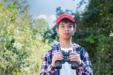Dürbün tutan Asyalı çocuk tropikal ormanda duruyor ve parkta vahşi hayvan görmek için bekliyor. Doğal ortamın tadını çıkarıyor ve yabani hayatı merakla izliyor..