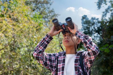 Asian boy holding binoculars and  standing in tropical forest waiting to see wildlife animals in local park. He is enjoying the natural surroundings and observing wildlife with curiosity. clipart