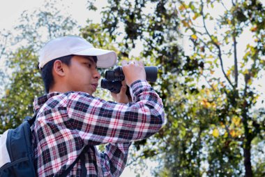 Dürbün tutan Asyalı çocuk tropikal ormanda duruyor ve parkta vahşi hayvan görmek için bekliyor. Doğal ortamın tadını çıkarıyor ve yabani hayatı merakla izliyor..