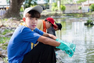 Asian young volunteers uniforms work together near a pond in their community by using nets to collect plastic bottles and other trash from the water to protect local spaces from pollution. clipart