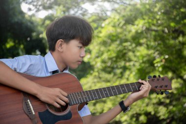 Okul üniformalı Asyalı bir okul çocuğu elinde ukulele, okul parkında oturuyor ve onu çalıyor..