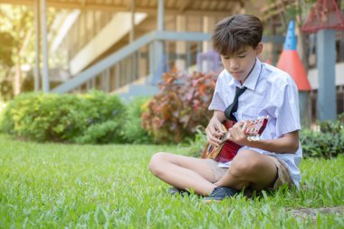 Okul üniformalı Asyalı bir okul çocuğu elinde ukulele, okul parkında oturuyor ve onu çalıyor..