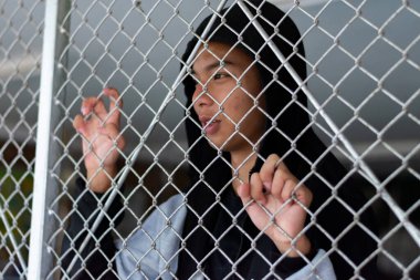 Gri kapüşonlu genç bir çocuk yerel bir ıslahevinde elleri metal bir çitin üzerinde duruyor, üzgün görünüyor ve özgürlüğünü istiyor..