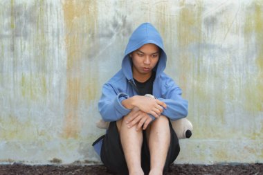 Mavi kapüşonlu Asyalı genç, oyuncak bebek tutuyor ve kirli duvarın önünde duruyor, üzgün görün, yumuşak odaklan, evsiz konsepti, hapsedilme ve gözaltı konsepti.