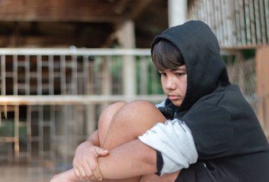 Şort giymiş, gri ve siyah kapüşonlu bir bornoz giymiş yüzü kasvetli ve üzgün bir şekilde kirli bir odada oturuyordu çünkü ailesinin sorunlarından ve cezasından kaçıyordu..