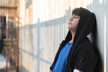 Şort giymiş, gri ve siyah kapüşonlu bir bornoz giymiş yüzü kasvetli ve üzgün bir şekilde kirli bir odada oturuyordu çünkü ailesinin sorunlarından ve cezasından kaçıyordu..