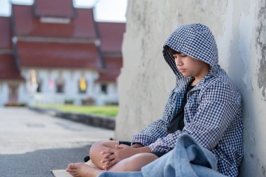 Koyu renk kapüşonlu ceket giyen Asyalı genç çocuk, taş bir çitin ya da bariyerin yanında düşünceli bir şekilde oturuyor. Arka planda çimento çitleri var, özünde, mutsuz, üzgün ruh hali kavramını vurguluyor..