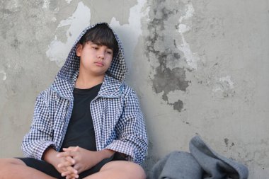 Siyah kapüşonlu Asyalı genç kirli zeminde oturuyor ve terk edilmiş bir binanın dağınık duvarında üzgün, yumuşak odaklı, evsiz konsepti, hapsedilmesi ve gözaltına alınması..