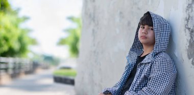 Koyu renk kapüşonlu ceket giyen Asyalı genç çocuk, taş bir çitin ya da bariyerin yanında düşünceli bir şekilde oturuyor. Arka planda çimento çitleri var, özünde, mutsuz, üzgün ruh hali kavramını vurguluyor..