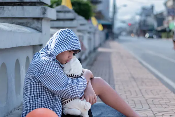 Koyu renk kapüşonlu ceket giyen Asyalı genç çocuk, taş bir çitin ya da bariyerin yanında düşünceli bir şekilde oturuyor. Arka planda çimento çitleri var, özünde, mutsuz, üzgün ruh hali kavramını vurguluyor..