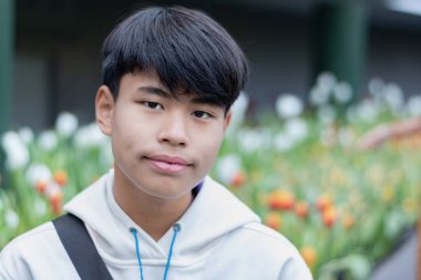 Beyaz kapüşonlu genç bir adam çiçek açan beyaz lalelerle çevrili bir bahçede oturuyor, sakin ve düşünceli bir ifadeyle, yemyeşil ve canlı çiçekler dingin ve ferahlatıcı bir atmosfer yaratıyor..