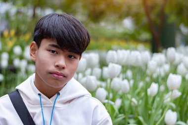 Beyaz kapüşonlu genç bir adam çiçek açan beyaz lalelerle çevrili bir bahçede oturuyor, sakin ve düşünceli bir ifadeyle, yemyeşil ve canlı çiçekler dingin ve ferahlatıcı bir atmosfer yaratıyor..