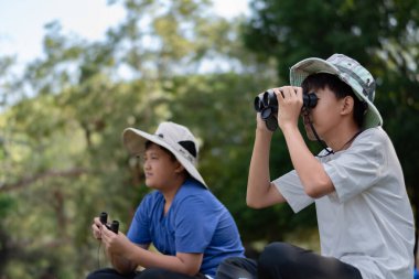 Doğayı keşfeden iki Asyalı çocuk, biri dürbün kullanıyor diğeri de not yazıyor. Yeşil, orman benzeri bir ortamda açık hava öğrenimi, merak ve çocukluk macerası..