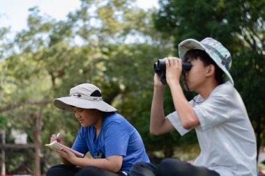 Doğayı keşfeden iki Asyalı çocuk, biri dürbün kullanıyor diğeri de not yazıyor. Yeşil, orman benzeri bir ortamda açık hava öğrenimi, merak ve çocukluk macerası..