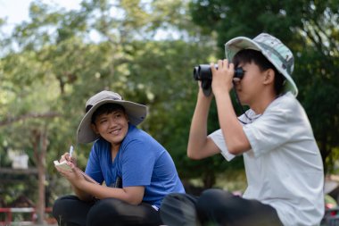 Doğayı keşfeden iki Asyalı çocuk, biri dürbün kullanıyor diğeri de not yazıyor. Yeşil, orman benzeri bir ortamda açık hava öğrenimi, merak ve çocukluk macerası..