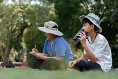 Doğayı keşfeden iki Asyalı çocuk, biri dürbün kullanıyor diğeri de not yazıyor. Yeşil, orman benzeri bir ortamda açık hava öğrenimi, merak ve çocukluk macerası..