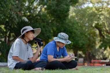 Doğayı keşfeden iki Asyalı çocuk, biri dürbün kullanıyor diğeri de not yazıyor. Yeşil, orman benzeri bir ortamda açık hava öğrenimi, merak ve çocukluk macerası..