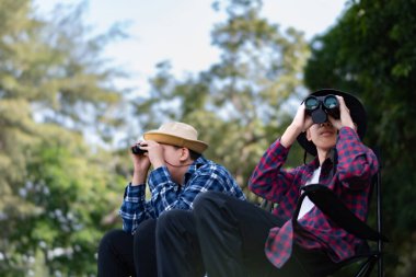 Doğayı keşfeden iki Asyalı çocuk, biri dürbün kullanıyor diğeri de not yazıyor. Yeşil, orman benzeri bir ortamda açık hava öğrenimi, merak ve çocukluk macerası..