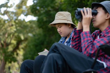 Doğayı keşfeden iki Asyalı çocuk, biri dürbün kullanıyor diğeri de not yazıyor. Yeşil, orman benzeri bir ortamda açık hava öğrenimi, merak ve çocukluk macerası..
