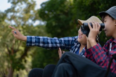 Doğayı keşfeden iki Asyalı çocuk, biri dürbün kullanıyor diğeri de not yazıyor. Yeşil, orman benzeri bir ortamda açık hava öğrenimi, merak ve çocukluk macerası..