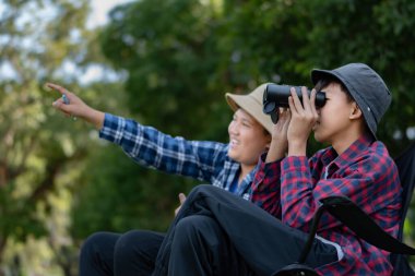 Doğayı keşfeden iki Asyalı çocuk, biri dürbün kullanıyor diğeri de not yazıyor. Yeşil, orman benzeri bir ortamda açık hava öğrenimi, merak ve çocukluk macerası..