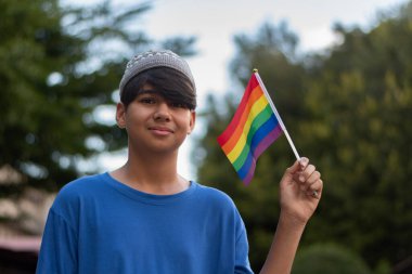 Açık havada dikilirken gökkuşağı LGBT gurur bayrağı tutan kendine güvenen genç bir çocuk. Gençlik eşitliği, çeşitliliği ve aşkı destekliyor. LGBTQ + pozitif ifade ve kapsayıcılığa sahip gurur ayı konsepti.